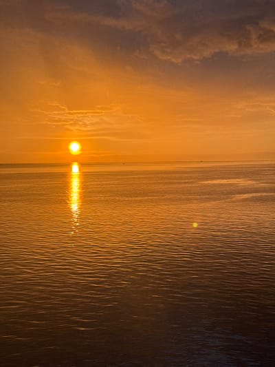 A serene sunset over the ocean is captured in this image. The sun, a bright orb of light, hovers just above the horizon, casting a golden glow across the sky and water. The sky is a canvas of warm hues, transitioning from a deep orange at the horizon to a softer, paler orange and then to a muted purple towards the top of the frame. Wispy clouds are scattered across the sky, catching the last rays of the sun.

The ocean surface is calm, with gentle ripples that reflect the sunlight, creating a shimmering path from the sun to the foreground. The water's surface is a warm, deep orange, reflecting the colors of the sky. In the distance, on the horizon, a faint silhouette of a small boat can be seen. The image conveys a sense of tranquility and the beauty of nature.

This scene does not depict any specific location within the city of Slobodka, Russia.
