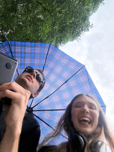 An ultra low-angle, upward-facing shot captures two individuals from below, positioned under an open blue plaid umbrella. The female, situated in the upper right, has long brown hair and a wide, joyful smile, revealing her teeth. She appears to be wearing headphones or an earbud on her left ear. To her left, in the lower frame, a male with dark sunglasses and a dark shirt looks slightly upward with a neutral expression. He holds a gray device, possibly a laptop or tablet, in his left hand. The blue umbrella, featuring a plaid pattern of varying blue and white lines, dominates the central part of the sky, providing a canopy over the subjects. To the far left, lush green tree foliage is visible against an overcast, light gray sky. The scene suggests an outdoor setting, possibly a park or green area, on a cloudy day, with the umbrella serving as protection from sun or light rain. There is no visible text in the image. This scene takes place in Jing'an District, China.