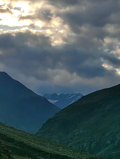 The image depicts a mountainous landscape under a dramatic sky filled with both dark, heavy clouds and bright, sunlit patches. The foreground features the steep, rocky slopes of mountains covered in sparse vegetation. Two prominent mountain ranges converge in the center of the frame, with the left range appearing darker and more shadowed, while the right range is bathed in a greener, more textured light. In the distance, further mountain peaks are visible, some with patches of snow. The sky is characterized by a gradient of dark blues and grays in the lower half, transitioning to brighter, white and yellow hues where the sun is breaking through the clouds. There are no people or man-made structures visible in the image, suggesting a remote, natural environment. The scene is consistent with the mountainous terrain found in regions like Damphug, India, known for its rugged topography and atmospheric skies.