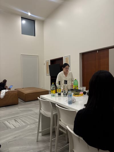 The photograph depicts an indoor scene, with two individuals partially visible. One person with long, dark hair is prominently in the foreground, obscuring much of the view. The second individual is situated behind a white table laden with bottles and containers, some of which appear to be alcoholic beverages. This second person is wearing a white hooded sweatshirt. White chairs surround the table. The floor is patterned with a grey, marble-like design. In the background, there is a dark shelf unit containing a television. The photograph was taken in Santiago Etla, Mexico.