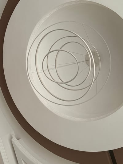 The scene depicts an upward-angled view within an interior architectural space, reportedly located in the city of Aragats, Armenia. A modern chandelier is centrally suspended, composed of five distinct, thin, circular rings of varying diameters. These rings, which appear to be light-colored or white, are arranged in an overlapping and descending configuration, supported by multiple fine suspension wires connected to a central point on the white ceiling above. The ceiling perimeter features an intricate, continuous decorative molding. A prominent, broad, dark brown curved band occupies the left and lower-left portions of the frame, indicating a spiraling architectural element, likely a staircase railing or a curved wall within an atrium or multi-story void. Additional white architectural details are visible beneath this brown band. No individuals, animals, or explicit actions are present within the frame.