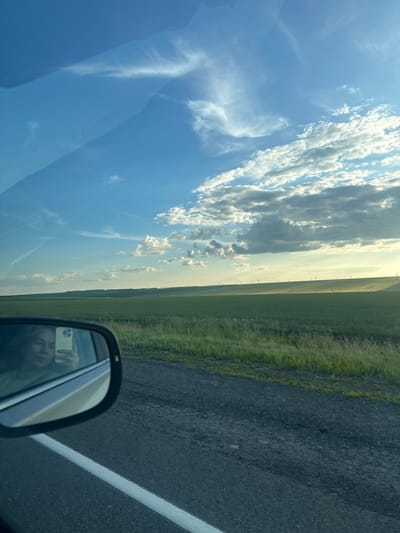 The image is a side view of a person taking a selfie in a car's side mirror. The mirror reflects a woman holding a smartphone, with a vast green field and a blue sky with scattered clouds in the background. The car is on a road, and the field stretches to the horizon where faint silhouettes of wind turbines are visible. The scene appears to be a rural landscape, possibly in the vicinity of Syzran, Russia, given the context provided. The lighting suggests it's either early morning or late afternoon, with the sun casting a warm glow over the landscape. The road surface is asphalt, with a white line marking the lane.