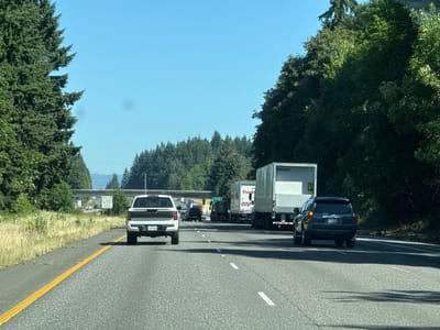 The image shows a highway scene on a clear day. In the foreground, a white pickup truck with license plate "KLW-6831" is driving in the right lane. To the right of the pickup truck, a dark colored SUV with license plate "BYW-3505" is in the same lane. Further ahead, there are several large trucks, including a white trailer truck with the number "24530492" visible and another trailer truck that appears to be from "Fred Meyer". The highway has multiple lanes and is bordered by tall green trees and vegetation. In the distance, there is an overpass with some vehicles on it, and signs indicating "EXIT 14". The sky is a bright, clear blue. The environment suggests a rural or suburban highway setting. The location is identified as Ridgefield, United States, based on the presence of "EXIT 14".