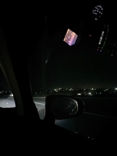 The image shows a view from inside a car at night, with the car driving on a road. In the foreground, a dark silhouette of the car's interior is visible, including part of the dashboard and a side mirror. The side mirror appears to be cracked. In the background, streetlights and illuminated buildings of a city are visible in the distance. The road ahead is lit by headlights. The location is identified as the city of Ciocîlteni, Moldova.