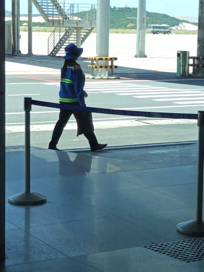 The image shows a view of Cam Ranh International Terminal in Bắc Cam Ranh, Vietnam. A person wearing a blue hat and a blue jacket with yellow stripes is walking. The person is wearing dark pants and dark shoes. A blue barrier tape stretches across the foreground. In the background, a bus is parked on the tarmac, and a set of stairs is visible. The ground appears to be tiled and reflective.