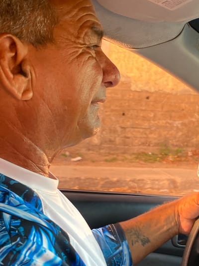 An older man is depicted in a medium shot, seen in profile within a vehicle, likely a car or truck, with the image rotated 90 degrees clockwise from its natural orientation. He has short, graying hair and a tanned, wrinkled complexion, particularly visible on his forehead and around his eyes. He is wearing a white collared shirt over a vibrant blue, black, and white patterned shirt. A dark, abstract tattoo is visible on his right forearm.

The man appears to be looking out a window, with a contemplative or observant expression. The background outside the vehicle, viewed through the window, is out of focus but shows a warm, earthy-toned wall or building, possibly made of stucco or brick, with hints of green and reddish foliage. The bright, warm lighting suggests it is daytime, possibly late afternoon or early morning, with direct sunlight illuminating the exterior. Parts of the car's interior, including a light-colored ceiling or pillar and a dark handle on the door frame, are visible. Faint, unreadable text can be seen on a label or sticker on the interior structure. The scene takes place in La Cruz del Pastel, Venezuela.