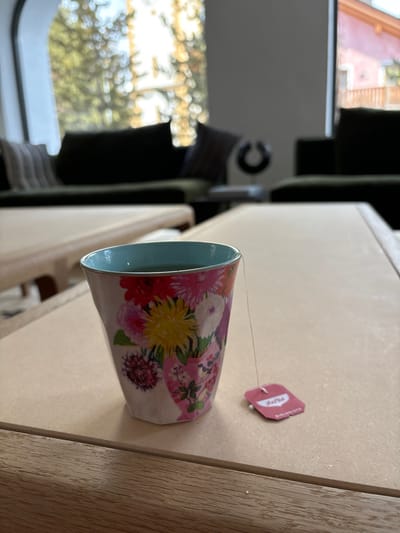 A floral teacup with a teabag sits on a wooden table. The teacup contains dark tea. The string from the teabag is attached to the cup, while the teabag tag lies on the table to the right of the cup. The table is light wood and appears to be the surface of a coffee table. The background shows a blurred, out-of-focus view of a living room. The living room is in a house in Celerina/Schlarigna, Switzerland, located at latitude 46.514 and longitude 9.859. Two large windows are in the background, one overlooking a wooded area and the other overlooking a neighboring house. The house is decorated with black and dark green couches.  The wooden table is made of light wood and the grain is visible.