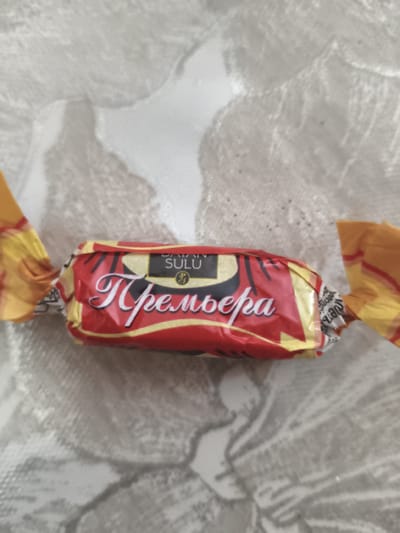 A single, red and yellow individually wrapped candy lies horizontally on a light gray, textured surface. The candy wrapper features the word "Премьера" in a prominent, stylized white script against a red background. Above this script, in smaller black text, is the word "SULU" with a circular emblem. The ends of the wrapper are twisted, revealing yellow and red patterned foil. The background surface appears to be a patterned tablecloth or placemat. The location is described as Lisakovsk, Kazakhstan.
