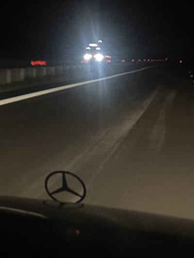 The image depicts a nighttime scene on a road, likely a highway, with a car's hood ornament, a Mercedes-Benz star, prominently in the foreground. The road has a white line marking its edge. In the distance, there are headlights of oncoming vehicles, some with red taillights visible, suggesting traffic. The overall lighting is dim, with the car's headlights creating bright glare. The image does not provide enough visual information to determine the specific location within the city of Zlatina, Bulgaria.