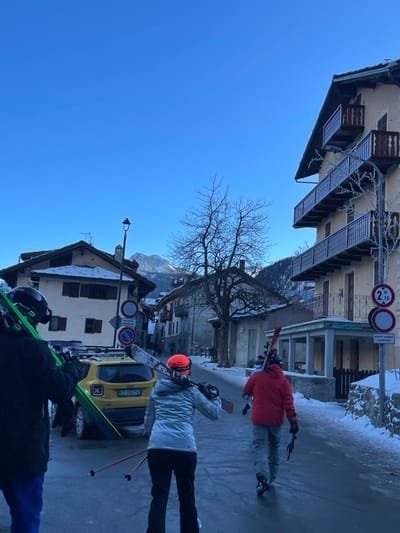 The image depicts a vibrant winter street scene in Courmayeur, Italy, under a clear, bright blue sky, suggesting daytime. Several individuals, likely skiers or snowboarders, are seen from behind or the side, walking along a street partially covered in snow and ice. They are dressed in winter attire, including jackets (red, light grey, dark) and helmets (orange, black), and are carrying skis with poles or snowboards.

The scene features traditional alpine architecture with multi-story buildings lining the street. Balconies on the buildings and the bare branches of a prominent tree in the middle ground are visibly frosted, indicating cold temperatures. In the background, snow-capped mountains are discernible. A yellow car, possibly a Fiat, with the Italian license plate "EZ 765YW" is parked on the side of the road.

Visible street signs include a circular white and red sign indicating a height restriction of "2.70 m," another circular red and white prohibitory sign, and a rectangular white sign with Italian text "eccetto residenti e proprietari" (except residents and owners). The ground exhibits patches of ice and snow reflecting the bright sky, contributing to the crisp, winter atmosphere. The activity suggests movement through a ski resort town, possibly to or from the slopes.