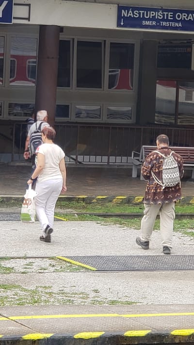 The image depicts a scene at the train station in Kraľovany, Slovakia. In the foreground, two individuals are walking away from the camera. The person on the left is a woman wearing white pants and a white t-shirt, carrying a white shopping bag. She has short, dark hair. To her right, a man is walking, wearing a patterned brown shirt, light-colored pants, and a patterned backpack. He has short, balding hair. In the background, there is a building with large windows reflecting a red train. A sign above the windows reads "NÁSTUPIŠTE ORAVA" and "SMER - TRSTENÁ". There are benches visible in the background, and the ground is a mix of gravel and concrete, with yellow and black safety lines. The overall environment suggests an outdoor public space, likely a train platform.