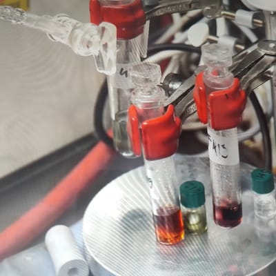 The image depicts a close-up view of laboratory equipment, specifically several glass vials containing liquids of varying colors, including amber, dark red, and clear. These vials are held in place by metal clamps with red silicone grips. One vial is labeled "F413". The vials are positioned on a textured metal surface, possibly a hot plate or a rack. In the background, there are other pieces of scientific apparatus, including clear plastic tubing and red-colored tubing. The overall scene suggests a chemical laboratory setting. The image does not contain any identifiable landmarks or elements that would allow for the determination of its location in Płock, Poland, or any other city.