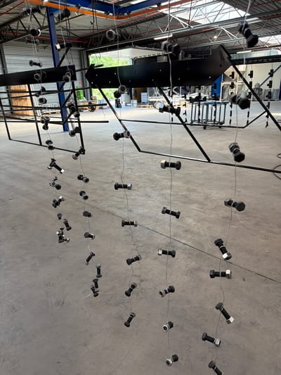 The image depicts an industrial setting, likely a workshop or manufacturing facility. Hanging from a black metal structure are numerous bolts and nuts, suspended by thin wires. These fasteners are arranged in vertical lines, creating a visually striking pattern. In the background, the facility's interior is visible, with industrial machinery, shelving units, and large overhead doors. The floor is a polished concrete surface. The presence of the bolts and nuts suggests they are either awaiting processing, such as painting or assembly, or are part of a decorative or artistic installation within the industrial space. The image was taken in Mol, Belgium.