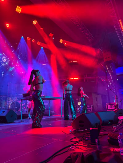 The image depicts a stage at an outdoor concert or performance in Akhalubani, Georgia. Three individuals, appearing to be female performers, are on stage. The performer in the foreground, facing away from the camera and to the right, wears a sequined, wide-legged jumpsuit and a halter top, with her hair in a ponytail. Another performer, also in a sequined jumpsuit and halter top, is further back on stage, facing left. A third performer, in a similar outfit, is visible to the right. The stage is lit with vibrant red and blue lights, creating a dramatic atmosphere. Stage equipment such as speakers, cables, a drum set, and a table with what appears to be DJ equipment are present. The background features a large screen displaying a graphic with a "G" logo and abstract shapes. The overall scene is one of a live entertainment event.