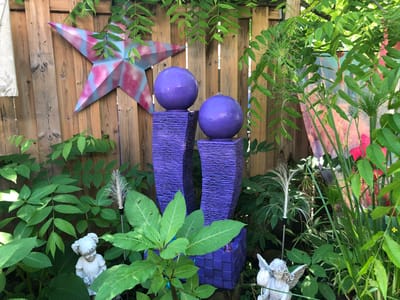 This image depicts a garden scene with various decorative elements and lush greenery. A wooden fence serves as the backdrop for several objects. A large, multi-colored star ornament, painted in shades of pink and blue, is affixed to the fence. In the center of the frame, a water feature is visible, consisting of two purple rectangular pedestals topped with purple spheres. A small amount of water appears to be gently flowing from the top sphere. The garden is populated with a variety of plants, including leafy green foliage and tall, reedy plants. Two small garden statues are present: one is a white cherub holding a flower, and the other is a white figurine of a child with pigtails. The lighting suggests it is daytime, with sunlight filtering through the leaves. The location is identified as a garden in the city of Severn, Canada.