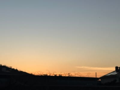 The image displays an outdoor scene in Piedrahíta, Spain, during twilight hours. The sky occupies the upper two-thirds of the frame, exhibiting a color gradient from light blue-grey at the top to warm orange-yellow at the horizon. A small, dark, irregular speck is visible in the upper right quadrant of the sky. The lower third of the image features silhouetted elements. On the left, an undulating dark landform, likely a hill, is present, topped with sparse, dark vegetation. Extending across the central midground is a horizontal dark barrier, resembling a wall or structure, topped with multiple coils of barbed or concertina wire. To the far right, a dark silhouette of a building, featuring a sloped roof and a chimney, is partially visible. The immediate foreground is a dark, undefined horizontal strip. No individuals are present in the scene. No discernible actions or interactions are occurring.