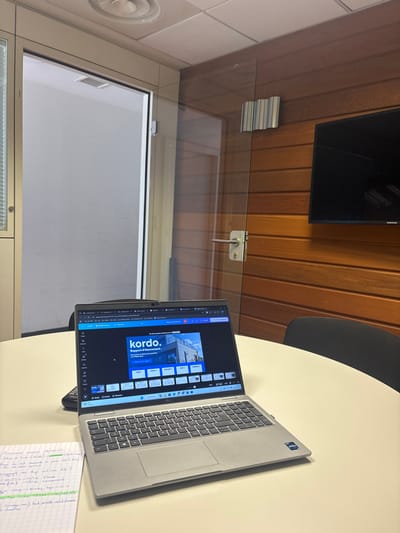 This image captures an indoor office or meeting room scene in the city of Lièpvre, France. A silver-gray laptop is open and positioned on a round, light-colored table, displaying a presentation or video editing interface featuring "kordo." and "Rapport d'étonnement" along with an image of a modern building and several slide thumbnails. A black calculator or similar device is partially visible to the left of the laptop, and an open spiral-bound notebook with handwritten French notes in blue and green ink occupies the bottom-left corner of the table.

The room's ceiling consists of white acoustic tiles with a circular vent. To the left, a glass door with a metallic frame opens into an adjacent white-walled space, with vertical blinds visible on the far left. To the right, a wall is covered with horizontal wooden slats. A clear glass partition with a metal door handle and lock separates this wooden wall section. A black Samsung flat-screen monitor is mounted on the wooden wall. A portion of a black office chair is visible behind the table on the right side. No individuals are present in the scene.
