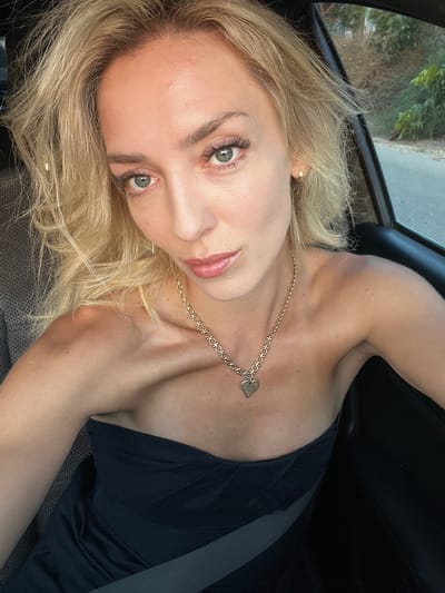 In a car, a blonde woman with light skin and blue eyes is taking a selfie. She has on a strapless black top and a gold necklace with a heart-shaped pendant. The woman's hair is messy and windblown. The background is blurred, but appears to be a street with trees and foliage, possibly in Beit Hanan, Israel.