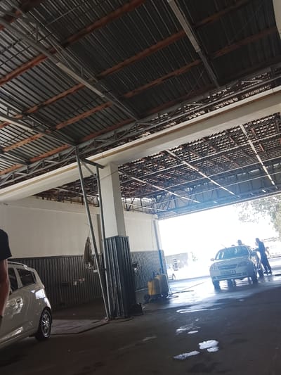 The image depicts an outdoor car wash facility in Kuk-Saray, Uzbekistan. The scene is characterized by a large, corrugated metal roof supported by a metal and concrete structure, creating a covered area for vehicle services. The floor is concrete and appears wet, with puddles and foam indicative of car washing activities.

In the foreground, a white compact car is parked. To the left, partially visible, is a person wearing a black shirt. Further back, a light-colored sedan is positioned in the car wash bay. Two individuals are interacting with this sedan; one is standing near the driver's side and appears to be working on the car, while another person is standing on the passenger side, possibly assisting. Another person, dressed in a dark blue shirt, is standing by the rear of the sedan. To the left of the sedan, a yellow piece of equipment on wheels, likely a pressure washer or foam dispenser, is present. In the background, a white vehicle is visible, along with further indistinct structures and trees. The overall lighting suggests it is daytime, with bright light emanating from the open end of the car wash.
