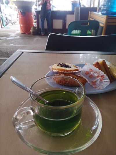 A table in an outdoor eatery in Palohlada, Indonesia, has a glass cup of green liquid and a plate of pastries. The cup is clear, with a handle, and rests on a clear saucer. A silver spoon is in the cup. The plate holds several pastries, including one with chocolate sprinkles and two pieces of bread. The table is light brown. In the background, a person in blue jeans and a pink shirt stands by a food stall. A large orange barrel is also visible.