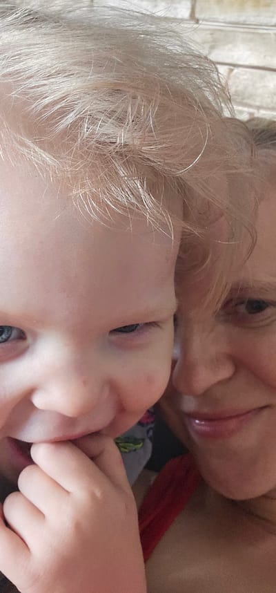 The image shows a close-up of two people, a woman and a child, lying down together. The woman's face is partially visible in the upper right, with her hair framing her face. She has blonde hair and appears to be smiling. The child is in the foreground, also with blonde hair. The child's face is turned towards the camera, with their eyes open and looking slightly to the left. Their hands are near their face, and one hand is touching the woman's face. The scene appears to be indoors, with soft lighting. The overall mood is intimate and affectionate. There is no visible text in the image. The location context provided is "Овощной, Russia," suggesting this is the general location where the photo was taken, though there are no specific visual cues in the image to confirm this. The image is oriented vertically, suggesting it might be a portrait-oriented photograph or a screenshot from a video.