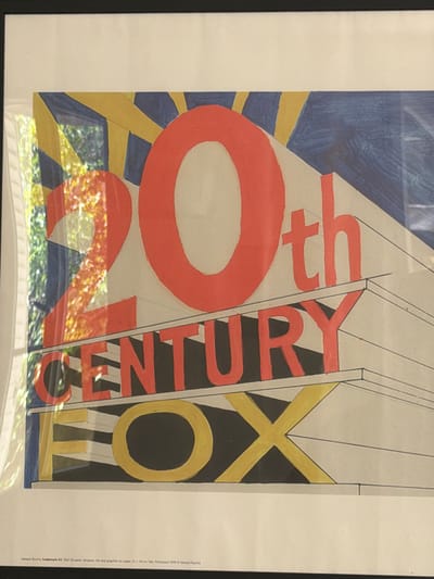 The image shows a framed print of an artwork depicting the 20th Century Fox logo. The text in the logo appears to be in red, black, and gold. The backdrop behind the logo consists of blue and yellow rays. The print is framed with a white border and a black frame. Text at the bottom indicates the artwork is "Edward Ruscha Trademark #5 1962 Oil paint, tempera, ink and graphite on paper 22 x 34 cm fate. Purchased 1999 © Edward Ruscha". The image also captures reflections of an outdoor environment, including foliage and possibly a building, indicating the print is photographed in an indoor setting. The location in the city of Aubervilliers, France, is not discernible from the visual information provided.
