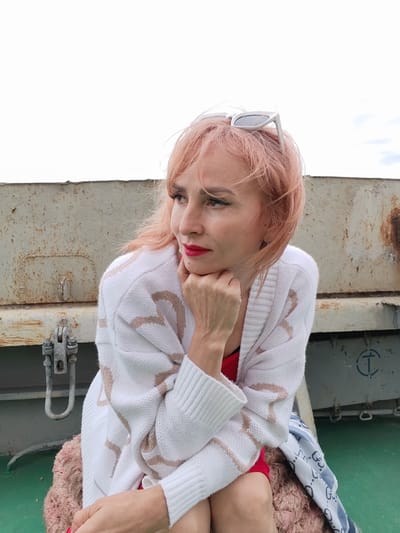 A woman with pink hair and red lipstick sits outdoors, resting her chin on her hand. She wears a white cardigan with a beige pattern and a red top. White sunglasses are perched on her head. She is seated on a fluffy pink cushion. In the background, there is a weathered, metal structure with rust stains and a green surface beneath. The sky is overcast. The location is identified as Bereg, Russia.