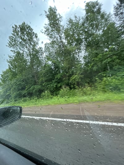 The image shows a view from inside a vehicle looking out through a rain-streaked windshield. The scene depicts a road with a white line marking the lane, bordered by lush green trees and vegetation. The sky is overcast, suggesting a rainy day. A side-view mirror of the vehicle is visible in the lower left corner of the frame. The location is identified as the city of Kulmas, Russia.