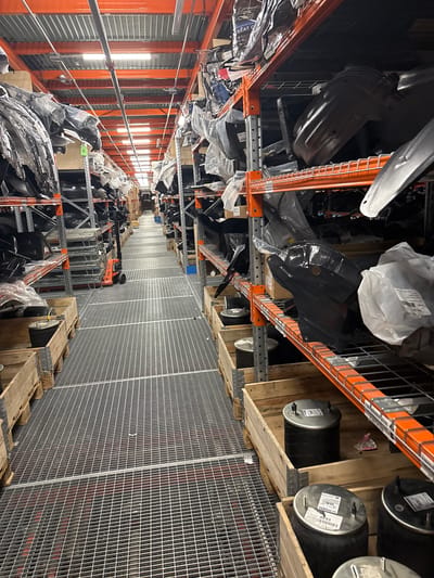 The image depicts the interior of a warehouse, likely located in an industrial area, although the city of Gara Elin Pelin, Bulgaria, cannot be conclusively determined from the visual information alone.
The scene is dominated by metal shelving units with orange frames. These shelves are densely packed with various items, many of which appear to be automotive parts, some wrapped in plastic. There are also black, cylindrical air brake chambers stored on pallets.
A wire mesh barrier extends along one side of the warehouse, possibly to secure a section or delineate a walkway. The lighting is provided by overhead fluorescent fixtures.