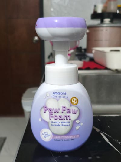 A light purple and white foam hand soap dispenser sits on a dark, reflective surface. The dispenser has a pump mechanism at the top and a rounded body with branding. Text on the body reads "Watsons", "LOVE MY SKIN", "Paw Paw Foam", "HAND WASH Lavender Scented", and "99.9% Antibacterial Tested Suitable For Sensitive Skin". A circular emblem with "3X PURIFYING" is also visible. The background is blurred, showing a brown door on the left and kitchen appliances and a sink area on the right. The overall impression is a product shot of a hand soap dispenser. The scene is indoors, likely in a home or retail environment. There are no people or animals present. The lighting is neutral. The location is unspecified.