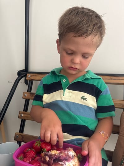 The image shows a young boy sitting on a wooden bench, looking down at a pink bowl of strawberries and dessert. The boy is wearing a striped polo shirt with green, blue, and white stripes. There are two temporary tattoos on his right arm, one appearing to be a cartoon character and the other with text. A white cup is partially visible to the left of the bowl. The background consists of a white wall and a dark metal structure. This scene does not provide any visual cues to suggest a specific location within the city of Les Rousses, France.