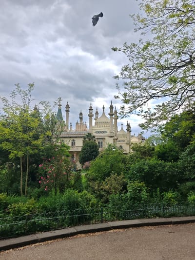 The image shows a view of the Royal Pavilion in Brighton, United Kingdom, seen through a dense array of greenery. The pavilion's ornate spires and domes rise above the trees and bushes. A bird is in flight in the upper center of the frame against an overcast sky. A black wrought iron fence runs along the lower part of the frame, separating the greenery from a paved pathway in the foreground.