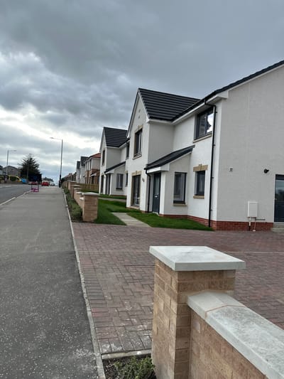The image depicts a residential street in Dunfermline, United Kingdom. A row of modern, two-story houses with white facades and dark-colored roofs lines the right side of the street. The houses feature dark-framed windows and doors. Small patches of green lawn separate the houses from the brick-paved walkway. The walkway is bordered by a concrete curb and a paved street. Streetlights illuminate the scene in the distance. Several vehicles are parked on the street further down. The sky is overcast with dark, heavy clouds. In the foreground is a low wall with brick pillars topped with marble slabs. No people are visible in the image.