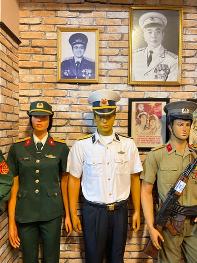 A display against a brick wall features mannequins dressed in various military uniforms. Portraits of uniformed figures hang on the wall next to the mannequins. One mannequin is wearing a tan uniform with a rifle, another is in a white naval uniform, and a third is dressed in a green uniform. A poster with Vietnamese text and images of soldiers is positioned between the top two mannequins. The overall impression is a historical or commemorative display related to the Vietnamese military.