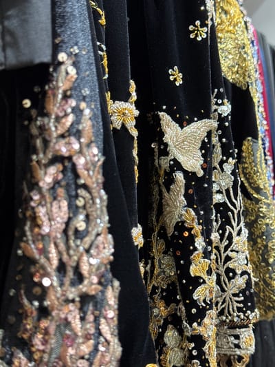 The image is a close-up of intricately embroidered black velvet garments, likely traditional clothing. The embroidery features metallic gold and silver threads, sequins, and beads, forming floral patterns and motifs such as butterflies and small star-like shapes. The lighting highlights the texture and shimmer of the embellishments. The garments are hanging, suggesting they are in a retail setting or a wardrobe. The background is dark, focusing attention on the detailed craftsmanship of the fabric. There are no people visible in the image, and the environment is not identifiable as being in Tanjungmeru, Indonesia.