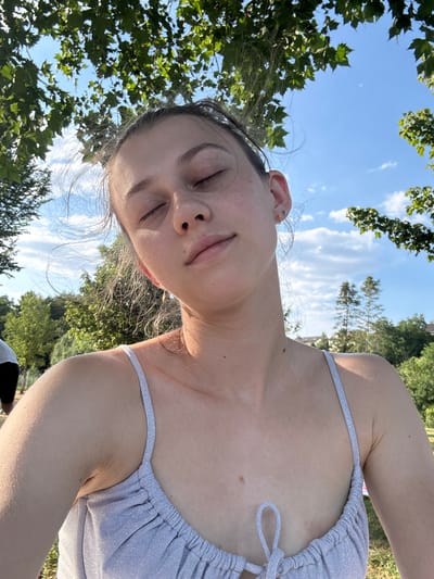 This is a tilted, close-up selfie of a young person lying down outdoors, with their head and upper body dominating the frame. The background consists of a clear blue sky with scattered white clouds, and lush green trees with dense foliage, suggesting a sunny day, possibly late afternoon or early evening due to the warm lighting on the skin.

The person is wearing a sparkling, light-colored halter top. Their eyes are closed, and their mouth is slightly open, giving them a relaxed or perhaps drowsy expression. Their hair is dark and appears slightly windblown, with some strands falling across their face. They have visible ear piercings.

The scene suggests a casual, leisurely activity, such as relaxing in a park or by a body of water on a warm day. The greenery and the lighting point to a summer or late spring setting. The orientation of the selfie and the overhead perspective imply the person is lying on their back and taking the photo with their arm extended upwards.

There is no visible text in the image. The location context of Bürgstadt, Germany, suggests this could be a scenic outdoor spot within that region.