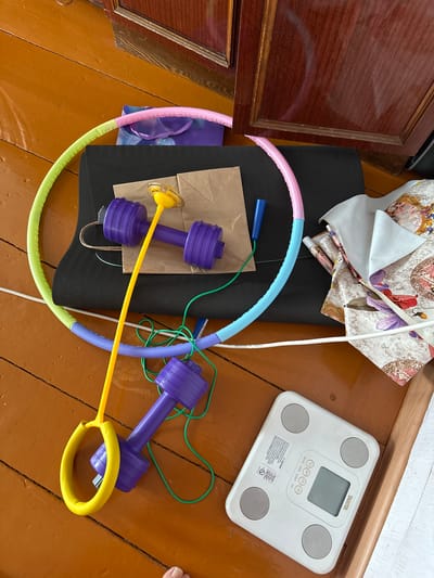 The image depicts a collection of fitness and personal care items scattered on a wooden floor. Prominently featured is a colorful hula hoop, segmented into yellow, light blue, and pink sections, resting atop a black yoga mat. Two purple toy dumbbells with yellow handles are also visible, one on the mat and another on the floor. A yellow jump rope with a ring is positioned across the dumbbells. A brown paper bag and some fabric with a floral pattern are present in the background. To the right, a white digital scale with a gray display and buttons is placed on the floor. The floor is made of light brown wooden planks. The scene suggests a domestic setting. The location is identified as Urazovo, Russia, though no specific contextual clues within the image confirm this.