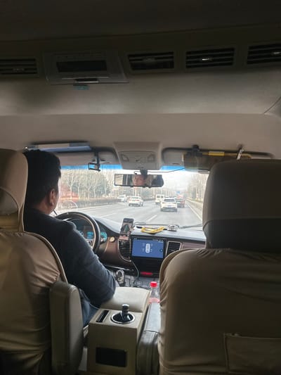 The image depicts the interior perspective from a rear seat of a moving vehicle, likely a van or SUV, traveling on a multi-lane road in Huangdao District, China. A male driver is visible from behind, wearing a dark garment and operating the vehicle with his hands on the steering wheel. His reflection, a male face looking forward, is visible in the wide rearview mirror.
The vehicle's dashboard features a large central display screen showing a white vehicle graphic, potentially indicating tire pressure or parking assist, and a smartphone mounted to the left, displaying a navigation interface. A gear selector knob is located on the center console, which also contains a cup holder holding a red-capped bottle. Overhead, air vents are visible, along with an overhead console unit and a dashcam mounted near the rearview mirror. A long, cylindrical object is secured above the passenger-side sun visor. The driver's seat and the seat in the immediate foreground are covered with light-colored fabric.
Outside the vehicle, a multi-lane highway is visible with moderate traffic, including several white and light-colored cars and a white van, all moving in the same direction. The roadside features a line of deciduous trees, currently bare, suggesting a colder season. The sky appears overcast or hazy, limiting visibility of the distant horizon.