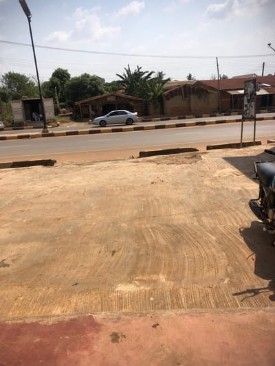 The image captures an outdoor street scene in Bada, Nigeria, under a clear sky with scattered white clouds. The foreground consists of a textured, tan-colored concrete or paved surface exhibiting semicircular finishing marks, with a reddish-brown section at the bottom.
A two-lane asphalt road, delineated by a median strip with brown and black painted curbs, runs horizontally across the midground. A silver sedan, featuring a rear spoiler, is observed in motion on the road, traveling from left to right.
To the left of the road, a tall black pole with a solar panel and LED light fixture stands. Behind it, a white shipping container, modified with an open side, reveals at least two individuals partially visible inside. Adjacent to the container, a wooden structure with a corrugated metal roof is present, followed by dense green trees and foliage.
On the right side of the road, several single-story buildings with corrugated metal roofs and unpainted walls are visible. A black rectangular sign on a pole stands on the right pavement. In the bottom right corner, a portion of a dark-colored motorcycle or scooter is visible, casting a shadow. Multiple power lines traverse the sky overhead.