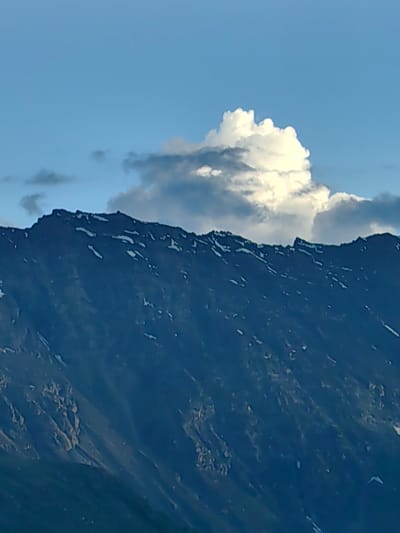 The image captures a majestic mountain range under a vast, clear blue sky dotted with white and gray clouds. The mountain's rugged slopes are covered in dark rock, with patches of snow and lighter-colored scree scattered across its surface, particularly along the jagged ridgeline. A prominent, brightly lit cumulus cloud formation billows above the mountain peak, suggesting a dynamic and possibly changing weather pattern. The lighting indicates it's likely daytime, with the sun illuminating the clouds and casting shadows on the mountain. The overall scene evokes a sense of natural grandeur and the raw beauty of a high-altitude environment. There are no people, objects, or visible text in the image. The setting is clearly mountainous and remote, fitting the provided location context of Kead, India, known for its mountainous terrain.