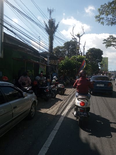 The image depicts a bustling street scene in Indonesia, identifiable by visible Indonesian license plates (B 3326 KBL and F 4788 WAL) and a political billboard for "PARTAI GOLKAR". This contradicts the stated location of Ban Phonhxavanh, Laos.

The scene is captured on a bright, sunny day with a partly cloudy sky. In the foreground, a person wearing a red jacket and a brown helmet is riding a white scooter away from the camera, occupying the right side of a paved road. Behind this scooter, a dark blue sedan is visible, also traveling in the same direction. To the left, a silver sedan is parked partially off the road, followed by a dirt/gravel shoulder where several motorcycles and scooters are parked. A group of approximately 5-7 individuals are standing and gathered near these parked vehicles and a one-story building with a green facade and an awning. Overhead, a dense network of electrical or communication wires spans across the frame. In the background, numerous trees are visible, including a tall, vine-covered tree topped with a cellular antenna tower, and a bare, gnarled tree further to the right. A large billboard displaying "PARTAI GOLKAR" and two male faces is situated further down the street on the right.