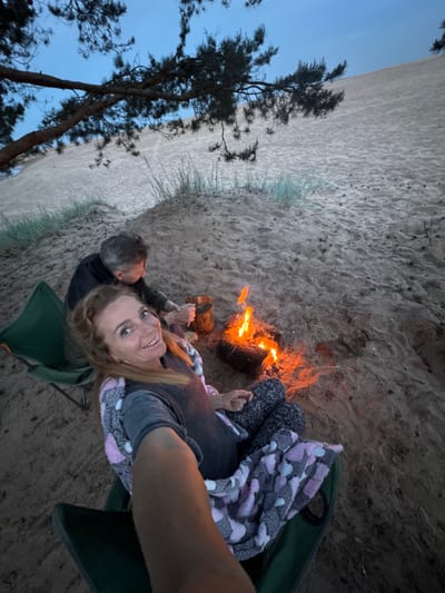 An overhead-angle selfie captures two individuals seated around a small campfire on a sandy dune in Bolshaya Izhora, Russia, during twilight. In the foreground, a woman with light brown hair, wearing a dark top, patterned pants, and a pink, grey, and white patterned blanket, smiles directly at the camera while holding the camera with her right arm, which is partially visible. She sits on a green folding camping chair. To her left and slightly behind, a man with short grey hair, wearing a dark jacket, sits on another green camping chair, facing away from the camera. He appears to be tending to a small pot or container placed next to a burning campfire. The campfire, centrally located, consists of a small log with visible orange flames. The immediate ground is sandy, scattered with sparse, light green vegetation. In the background, a large, expansive sand dune slopes upwards under a pale blue sky, with dark, pine-like tree branches extending from the upper left and right across the frame.