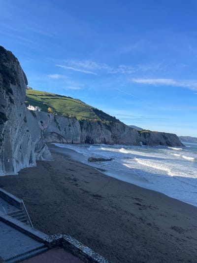 The image depicts Itzurun Beach in Zumaia, Spain, under a clear blue sky with sparse, wispy clouds and a single contrail. On the left, massive grey and brown stratified rock formations, characteristic of flysch geology, rise sharply from the dark sand beach. Green grassy hills, dotted with at least two small white houses, cap these cliffs. The beach extends into the mid-ground, where the Atlantic Ocean displays white-capped waves breaking towards the shore, casting elongated shadows inland. Several dark rocks are visible in the shallow water near the beach. In the lower-left foreground, a grey stone and concrete staircase with a retaining wall descends towards the beach, partially obscuring the sand. Footprints are discernible on the dark sand. No individuals are present in the scene.