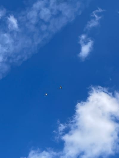 The image depicts a bright blue sky with scattered white clouds. Two small, light-colored airplanes are visible in the sky. One airplane is positioned to the left and lower than the other. The airplane on the right appears to be slightly larger in the frame, indicating it may be closer or larger. The clouds are wispy and diffuse, with some larger, more defined formations in the lower right portion of the image. The scene appears to be taken from the ground looking upwards. The image does not provide any specific details or visual cues that would allow for the identification of a location within Röllshausen, Germany.