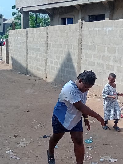 The image depicts a daytime scene on a dirt path in Ifite Awka, Nigeria. In the foreground, an adult individual with dark skin and dreadlocks wears a light blue and white short-sleeved shirt, a dark skirt or shorts, and dark Crocs-style footwear. This individual is positioned with their body angled to the left, leaning forward, and their right hand extended downwards towards the ground, appearing to be in motion or engaged in an action near scattered debris on the path. A yellow bracelet is visible on the individual's right wrist.
Behind and to the right of the adult stands a child, also with dark skin, wearing a white patterned long-sleeved dress or tunic over dark pants and open-toed sandals. The child is looking towards the adult.
The ground is a light brown, uneven dirt surface with scattered litter, including paper fragments, plastic pieces, and other small debris.
In the mid-ground, a gray concrete block wall extends horizontally across the frame. Above this wall, an unfinished or unpainted building is visible, featuring exposed concrete support structures, a balcony area, and two windows; one window has blue-tinted glass, and the other appears dark. A large, dark, triangular shadow from an unseen object is cast upon the concrete block wall. In the far background, additional structures and green foliage are discernible. To the far left, items of clothing are draped over a low structure or fence.