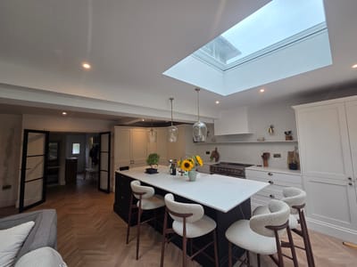This image depicts a modern, well-lit kitchen and dining area, likely in a residential setting in Elmbridge, United Kingdom. The scene is dominated by a large kitchen island with a white countertop, featuring several stylish, cream-colored bar stools. On the island, a vase of sunflowers adds a touch of natural beauty.
The kitchen itself is spacious and features contemporary cabinetry in both dark and light finishes. A sleek range cooker is visible, suggesting a functional and well-equipped cooking space. Above the island, decorative pendant lights hang, adding to the ambiance.
A prominent feature of the space is a large skylight in the ceiling, which floods the area with natural light, indicating it is daytime. The flooring is a distinctive herringbone pattern in a warm wood tone. In the background, a doorway with black-framed glass doors leads into another room, revealing hints of furniture and a wooden bench.
The overall impression is one of modern elegance and comfort, with a focus on natural light and stylish design. There are no people visible in the image, and no text is present. The scene suggests a recently completed renovation or a new build, emphasizing a clean and sophisticated aesthetic.