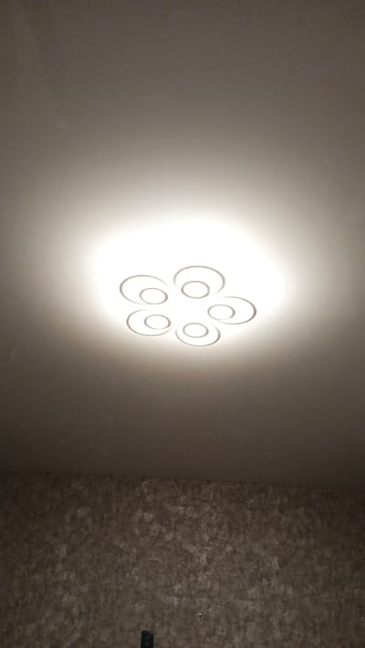 The image shows a ceiling light fixture with a modern, circular design. The fixture is illuminated, casting a bright glow on the ceiling. The light is composed of six concentric rings arranged in a flower-like pattern. Below the ceiling, a textured wall is visible, with a patterned wallpaper in shades of brown and grey. The lighting suggests that it is likely nighttime or indoors with artificial lighting. There is no visible text in the image. The location is noted as Gulkevichi, Russia.
