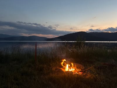 A campfire burns brightly in the foreground on a grassy shore, casting a warm glow on the surrounding dry grass and a few rocks. Tall reeds and grasses grow along the water's edge. Beyond the shore, a large body of water stretches out, with gentle ripples reflecting the twilight sky. In the distance, silhouetted hills form the horizon, their dark forms contrasting with the soft hues of the sky. The sky itself is a gradient of soft blues and pale oranges, with a few scattered clouds tinged with pink and gray. A single bright star is visible in the upper right portion of the sky. The scene evokes a sense of peace and tranquility, suggestive of an evening spent camping or enjoying a quiet moment by the water. The time of day is dusk or early evening. There is no visible text in the image. The location context is Batak, Bulgaria.