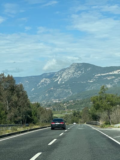 The image captures a highway scene in the Ula district of Muğla Province, Turkey, under a partially cloudy blue sky. The perspective is from a moving vehicle, looking forward down a two-lane asphalt road. White dashed lines mark the center of the road, and solid white lines delineate the shoulders. A black sedan, likely a Honda Civic, is positioned in the foreground, traveling away from the viewer in the right lane; its rear license plate begins with the provincial code "48". Further down the road, a white vehicle is also visible, receding into the distance.

On the left side of the road, a metal guardrail separates the pavement from dense green and brown trees. On the right, sparse vegetation lines an embankment before meeting more distant trees and a bridge structure. The mid-ground reveals a valley containing a cluster of residential buildings, indicating a settlement. Dominating the background are large mountains, covered extensively with green coniferous vegetation and featuring prominent grey rock formations. The highest visible mountain peak is topped with multiple antenna or communication tower structures. The scene is illuminated by natural daylight.