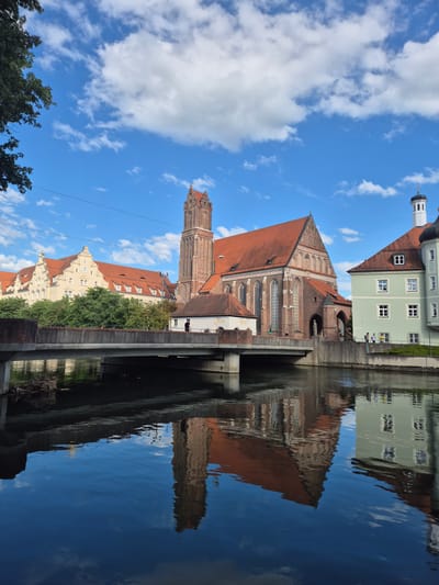 The image depicts a serene canal scene in Landshut, Germany, dominated by a majestic Gothic brick church with a tall tower, its orange-tiled roof gleaming under a partly cloudy blue sky. To the left, a pale yellow building with a similar red roof and a clock face is visible. On the right, a pastel green building with white-framed windows and a dark turret stands adjacent to the church. A concrete bridge spans the canal in the foreground, with the reflections of the buildings, bridge, and sky creating a mirrored image on the calm water. A few people are discernible on the canal bank and crossing the bridge, appearing as small figures engaged in casual strolling. The overall environment suggests a peaceful urban setting with a strong sense of historical architecture.